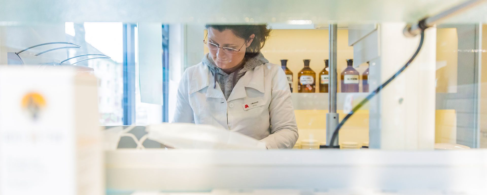 Apothekenmitarbeiterin in weißem Kittel arbeitet hinter dem Tresen, im Hintergrund stehen braune Apothekenflaschen in Regalen.