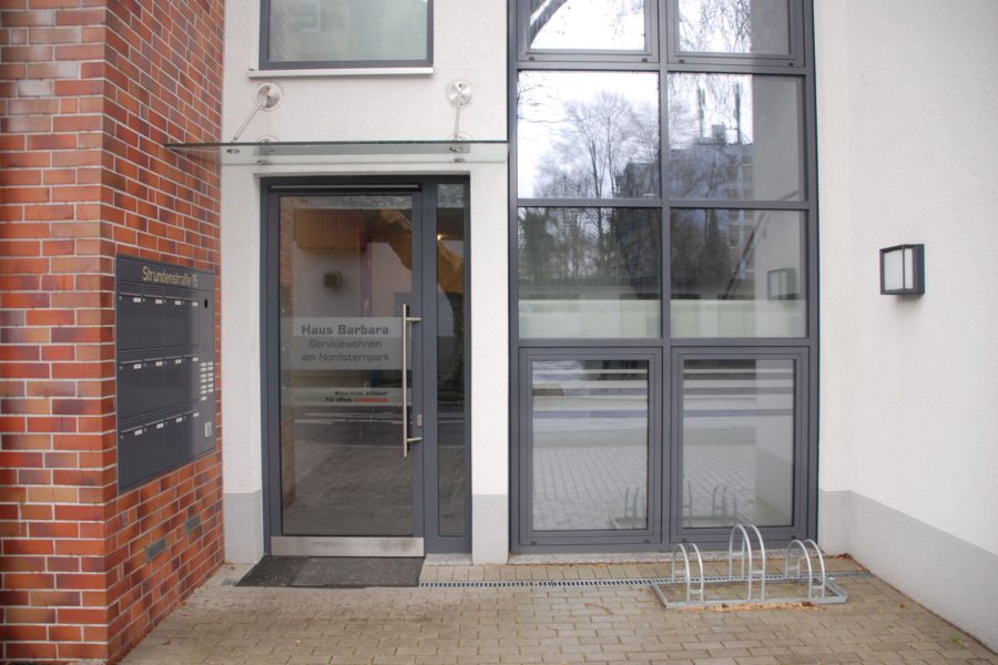 Eingangsbereich eines modernen Gebäudes mit Glastür, großen Fenstern und Briefkästen an der Wand