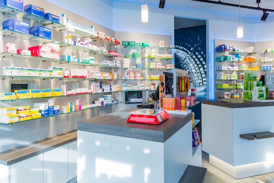 Innenansicht einer modernen Apotheke mit beleuchteten Regalen voller Medikamente und Verkaufsdisplays an der Theke.