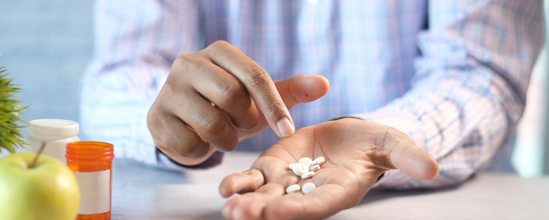 Eine Person hält mehrere weiße Tabletten in der Hand und zeigt mit dem Finger darauf; daneben stehen Medikamentenflaschen und ein grüner Apfel auf dem Tisch.